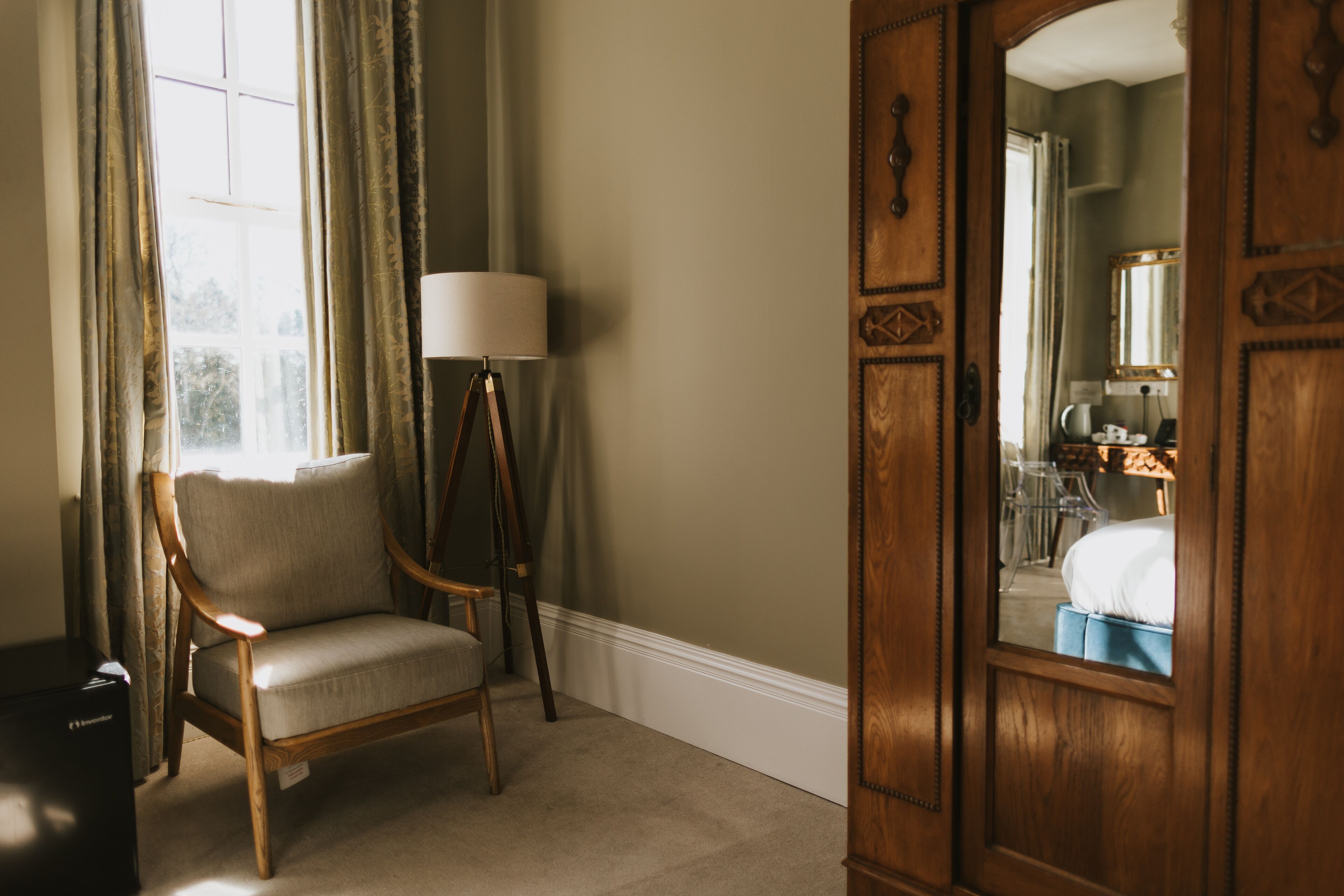 A room with a wooden chair, a floor lamp, and a large mirror reflecting a bed and bathroom.
