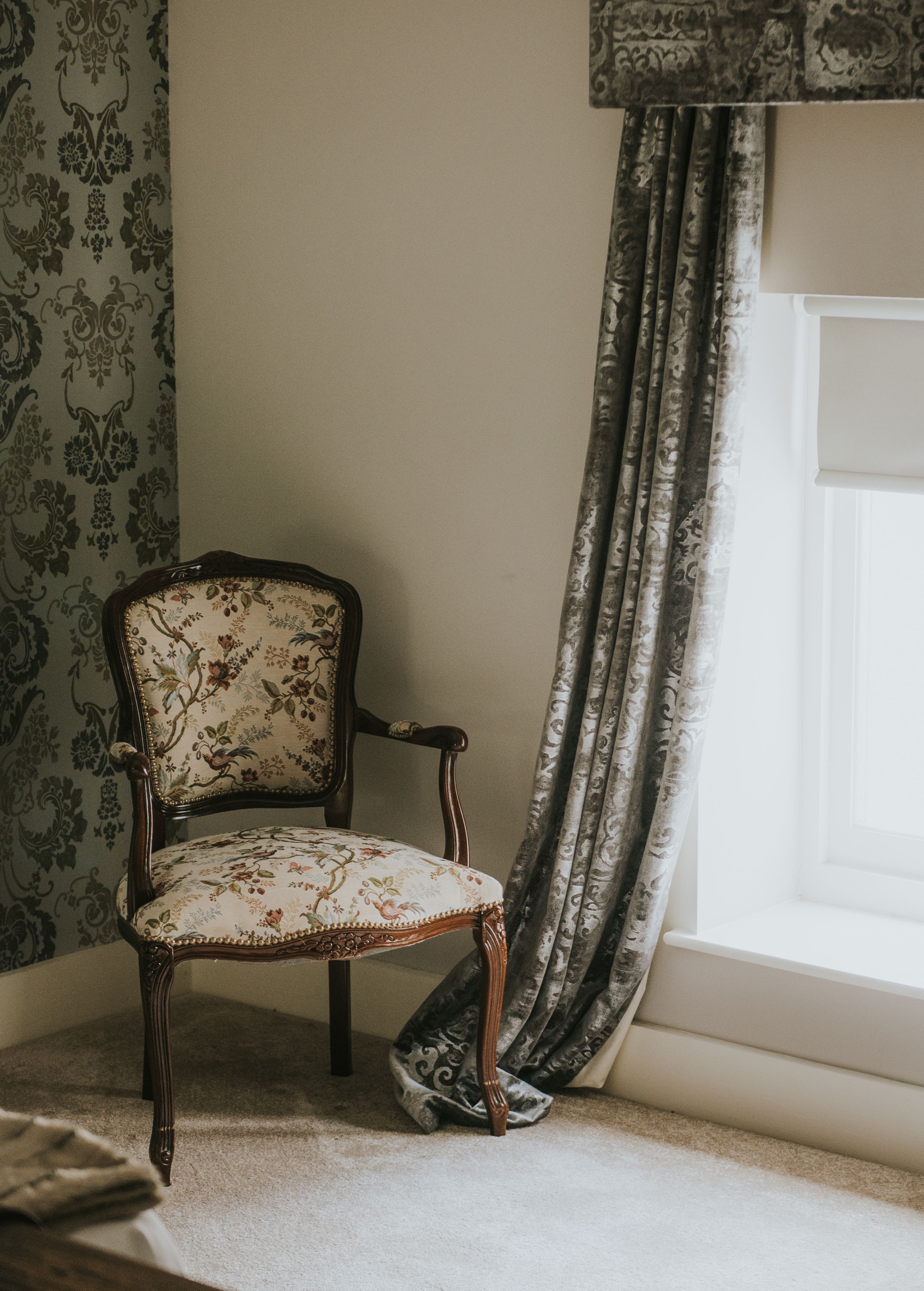 A corner of a room with a patterned wallpaper, a wooden chair with floral upholstery, and a window with a curtain.