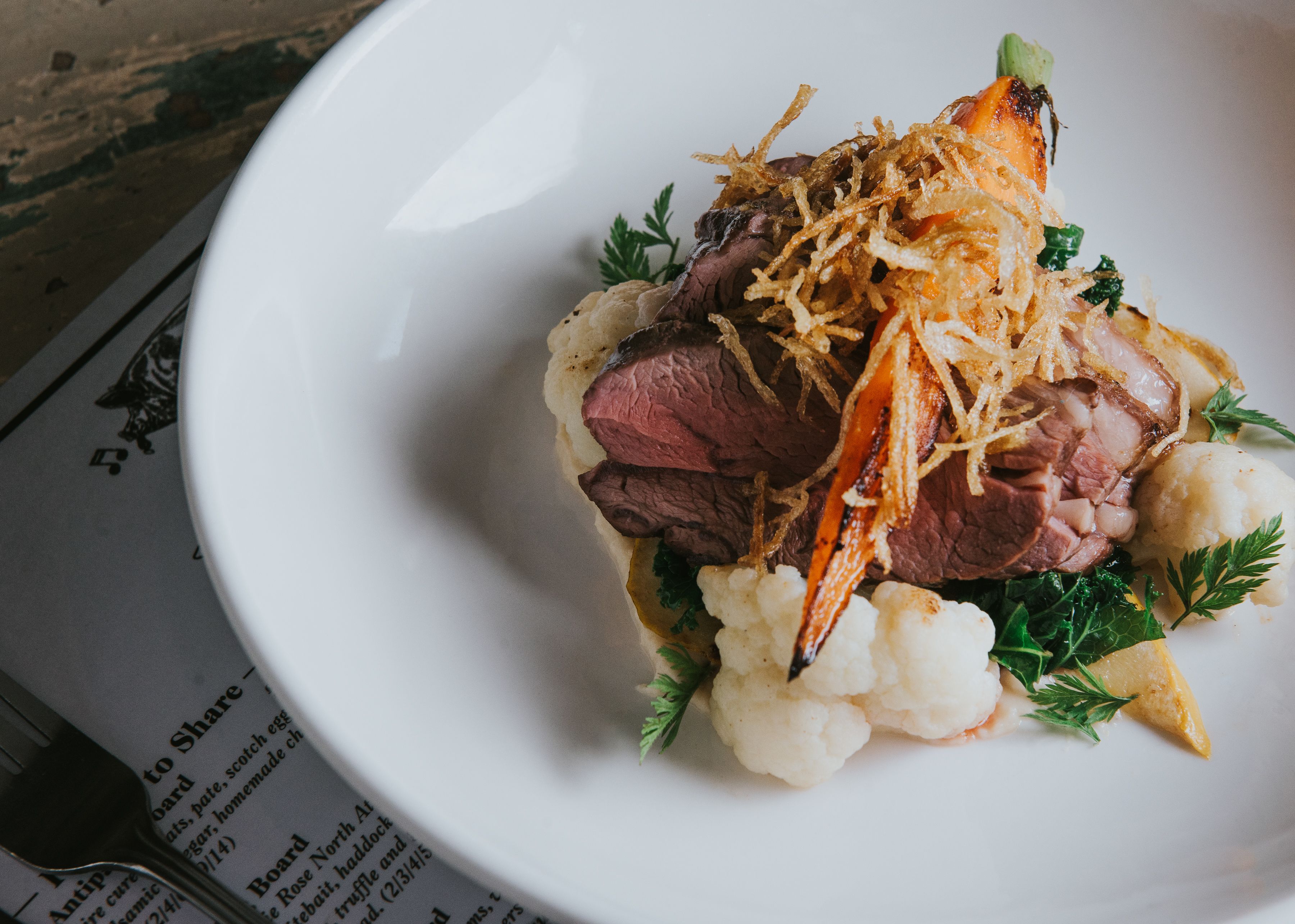A white plate with a dish of food, including meat and vegetables, on a table with a menu underneath.