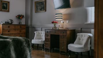 A room with a wooden dresser, two white armchairs, a desk with a lamp, a television mounted on the wall, and a window with blinds.