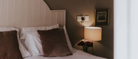 A neatly made bed with white and brown pillows, a wall-mounted lamp, and a framed picture on the wall.
