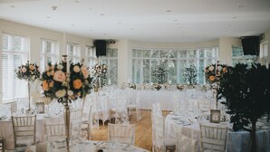 Uma sala de banquetes com mesas redondas cobertas por toalhas brancas, decorada com arranjos florais e castiçais.