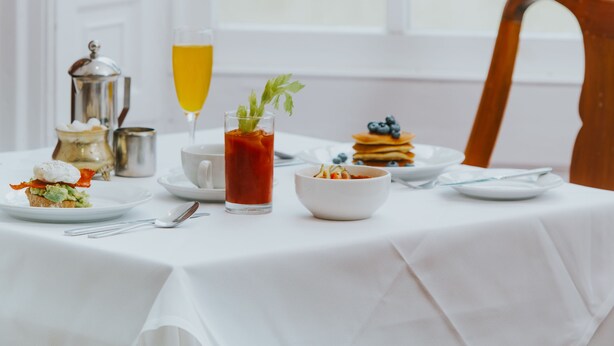 Mesa de café da manhã com diversos pratos, incluindo panquecas e um copo de suco de laranja, sobre uma toalha de mesa branca, com vista para o verde através de uma janela.
