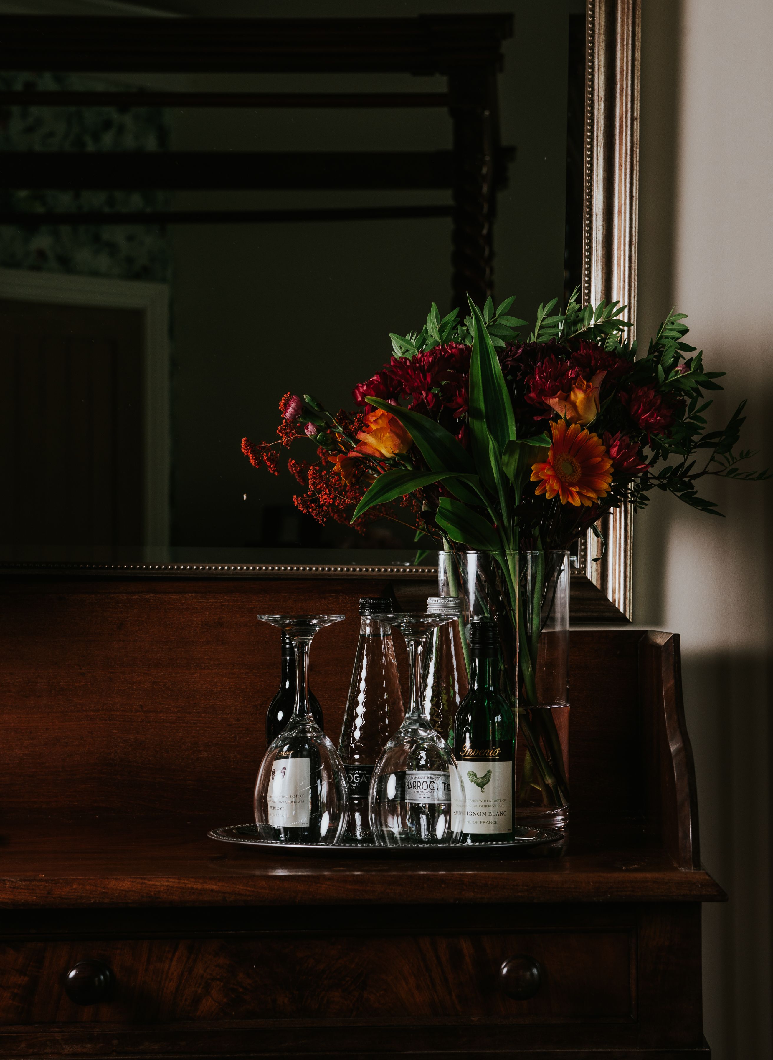 A wooden dresser with a mirror, a vase of flowers, and several wine bottles.
