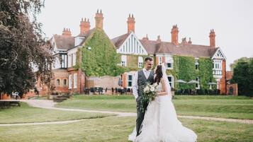 A couple in a wedding dress and suit standing in front of a large, ivy-covered building with a grassy lawn.