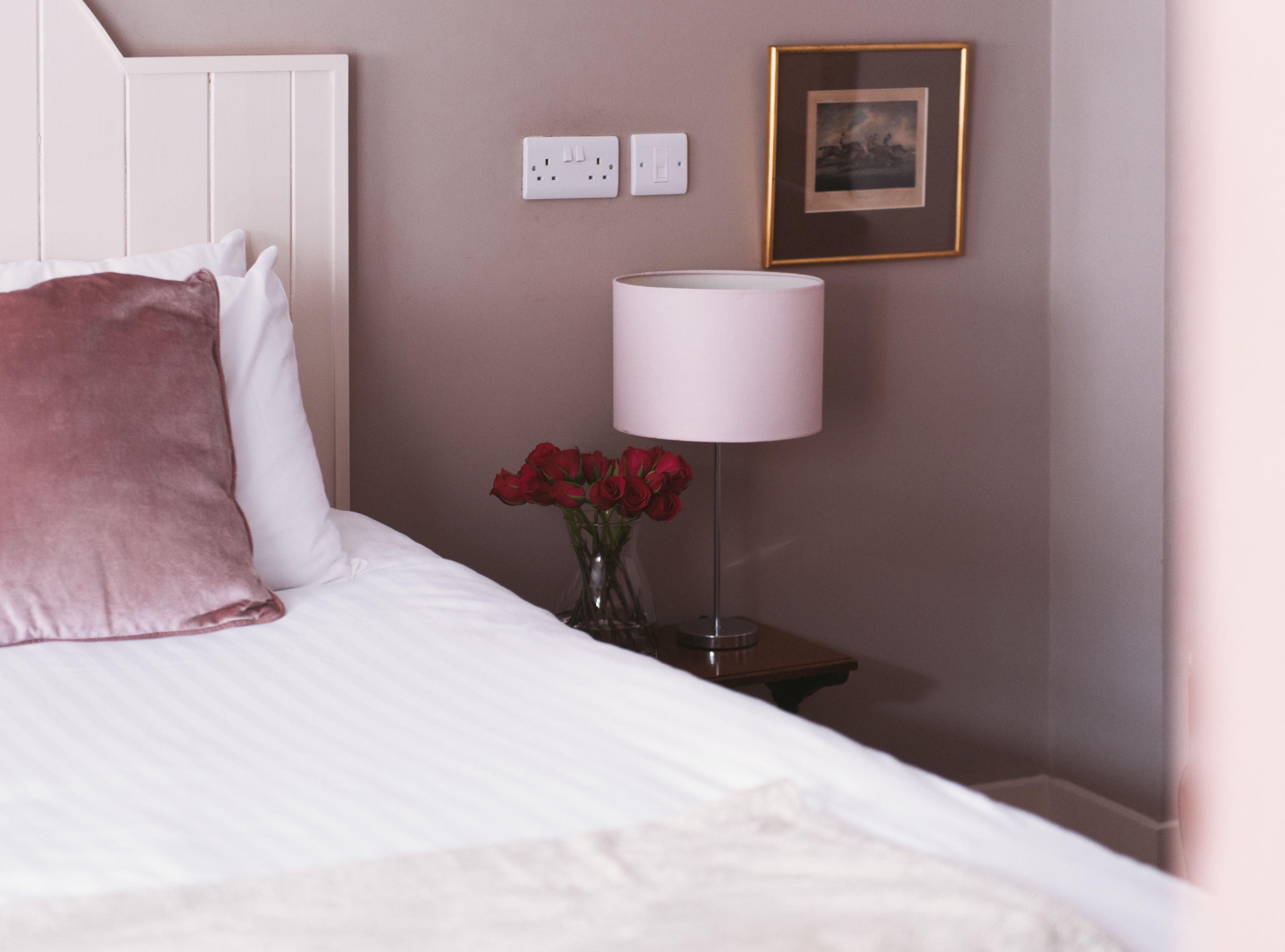 Una cama bien hecha con almohadas blancas y rosas, una mesita de noche con una lámpara y un jarrón con rosas rojas.