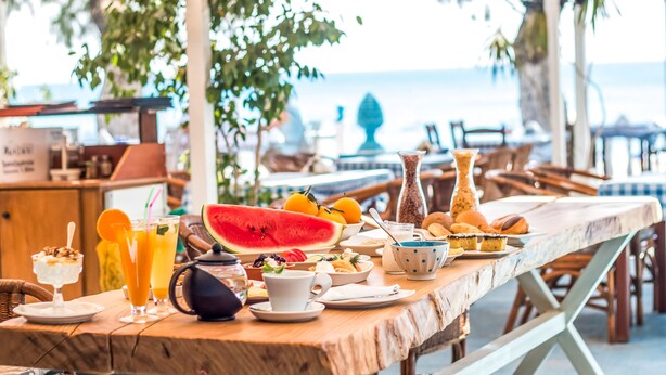 Ein Holztisch mit verschiedenen Frühstücksleckereien, darunter Früchte, Gebäck und Getränke, draußen mit Blick auf das Meer.