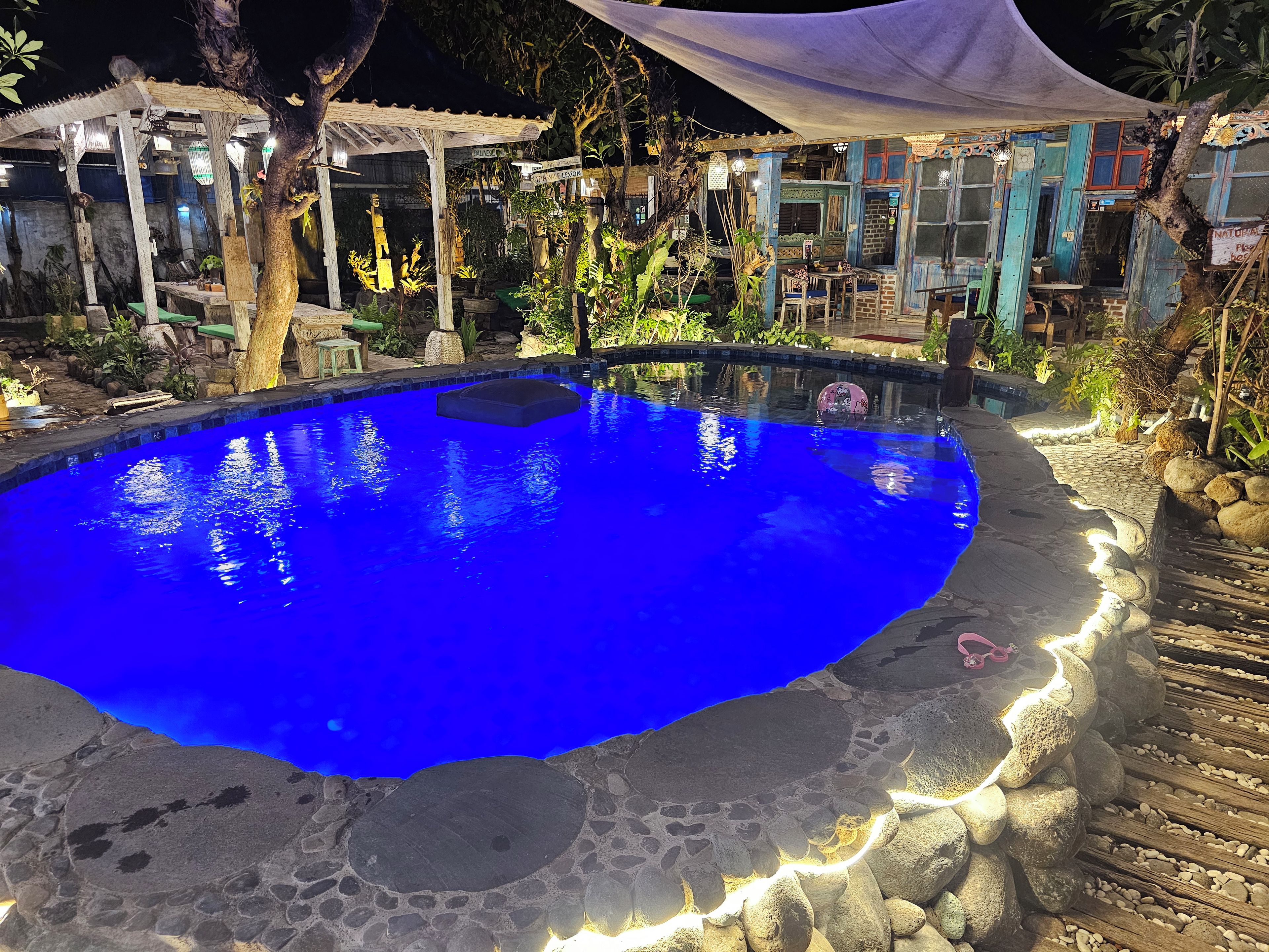 A swimming pool with a stone border, surrounded by a garden with potted plants and a gazebo.