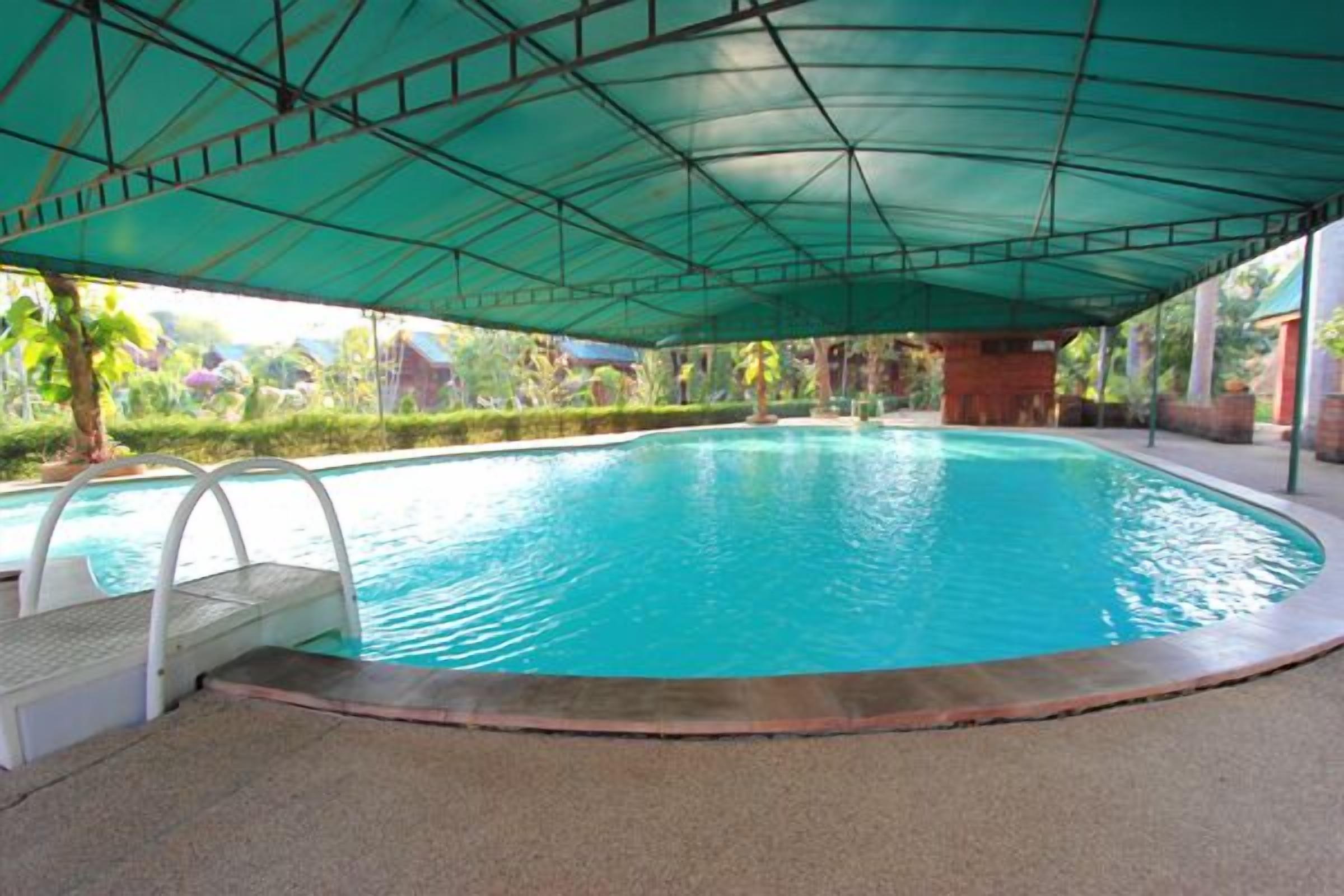 A swimming pool with a covered area, a diving board, and a poolside area with a view of greenery.