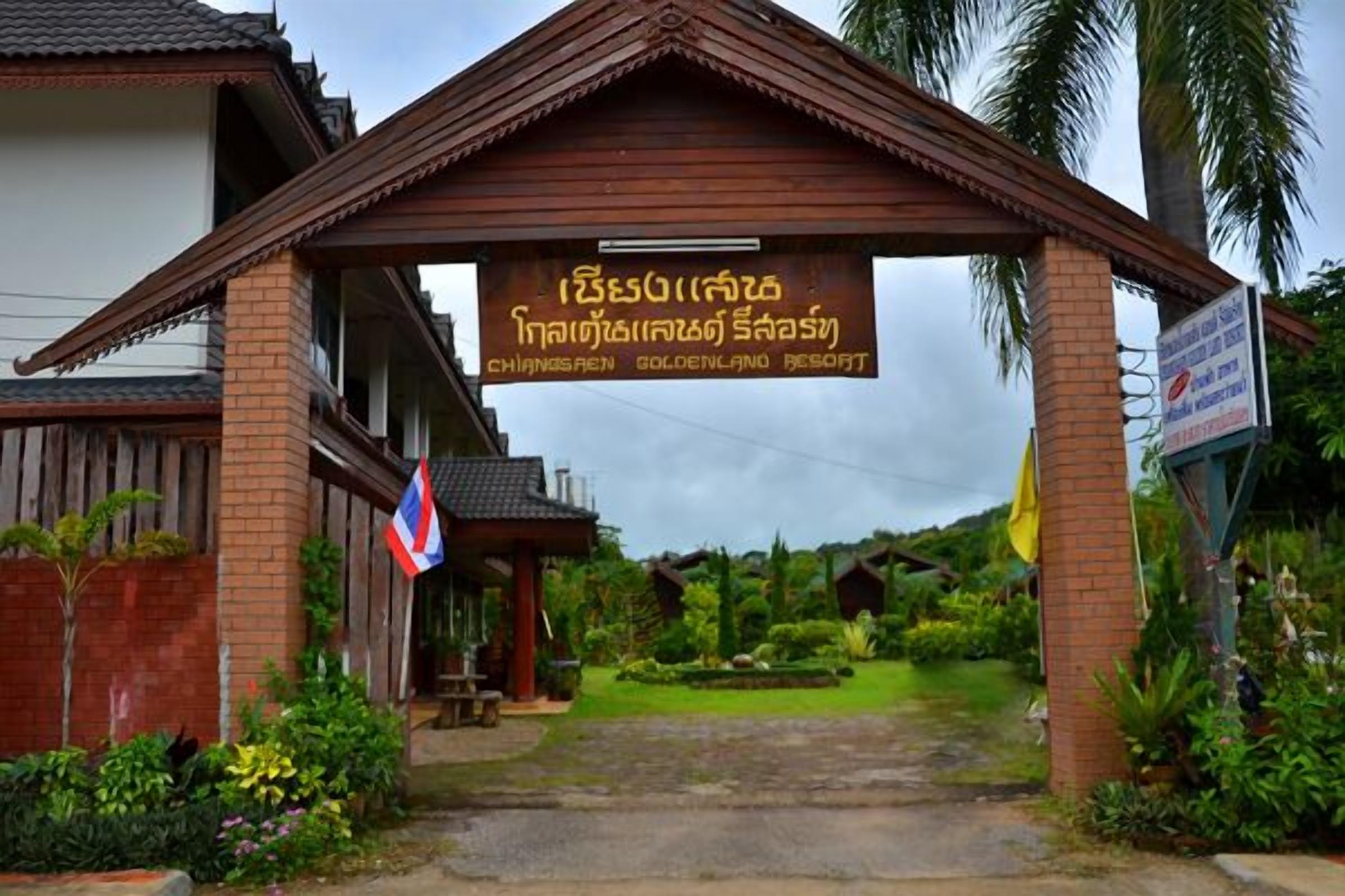 The entrance to a resort with a sign in a local language, surrounded by greenery and a traditional architectural style.