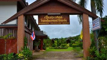 The entrance to a resort with a sign in a local language, surrounded by greenery and a traditional architectural style.