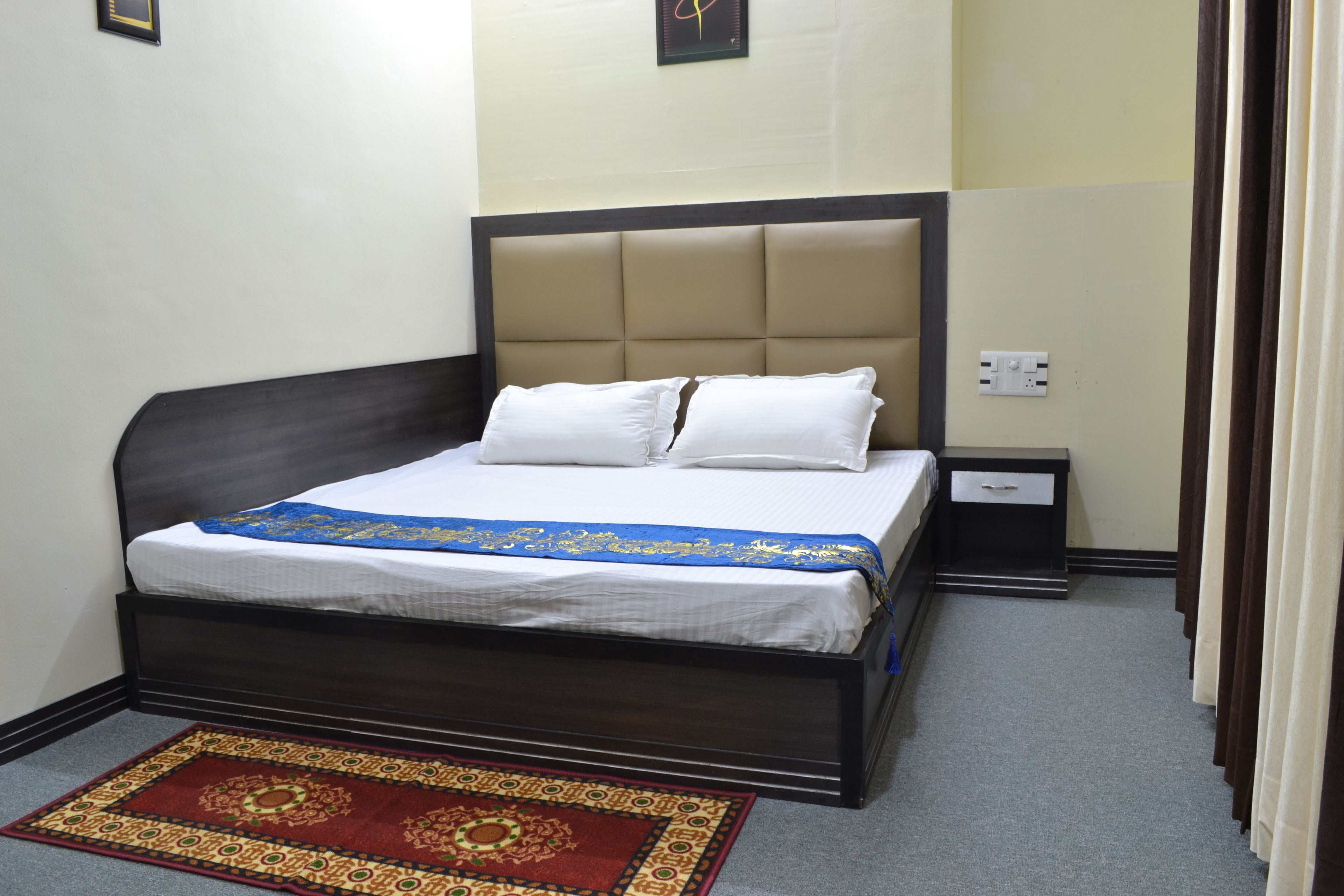 A neatly made bed with a headboard, a bedside table with a lamp, and a red patterned rug on the floor.