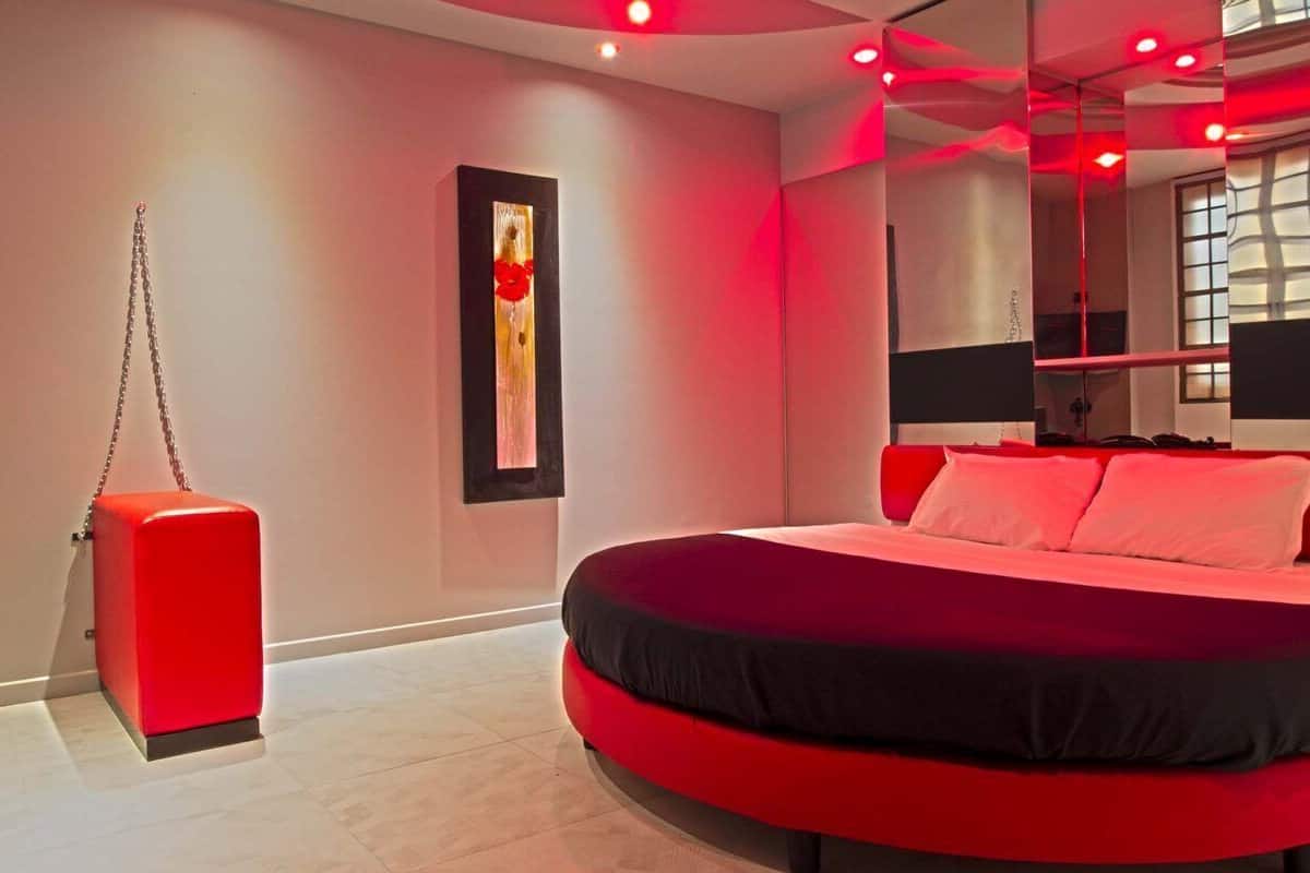 A modern bedroom with a large bed, a red accent wall, and a unique window design.