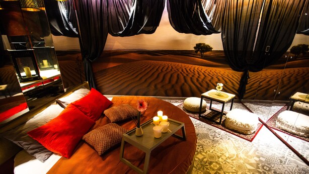A room with a curved bed, red pillows, a small table with candles, and a view of a desert landscape through sheer curtains.