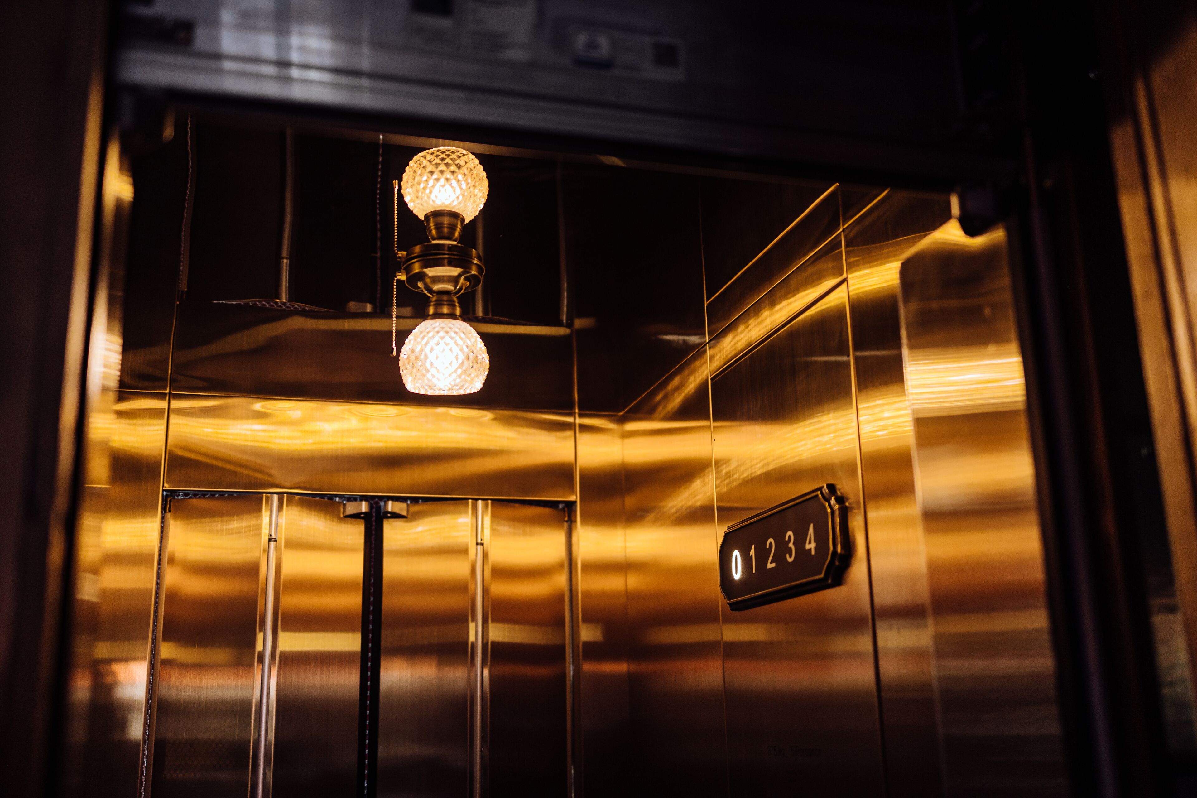 Interior de elevador com acabamento metálico, luminária e número de elevador visível.