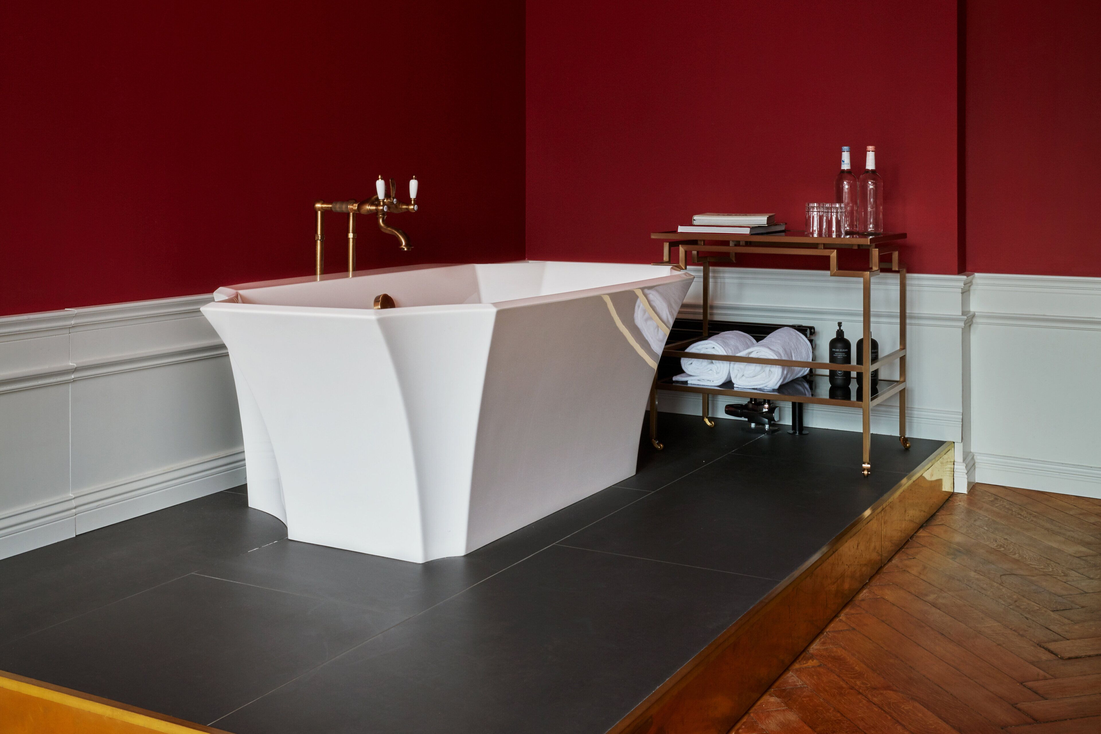 Une salle de bain moderne avec une baignoire sur pieds, une étagère en bois où sont rangés les articles de toilette et un mur d’accent rouge.