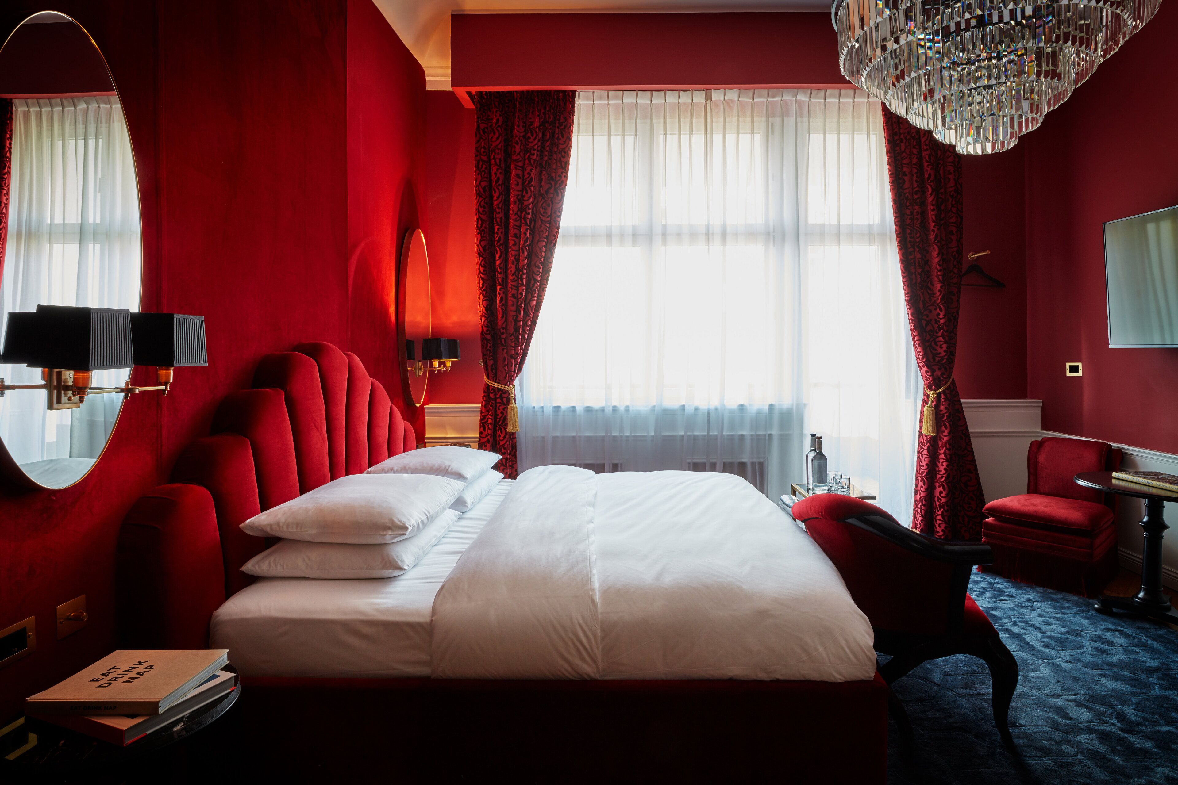 A hotel room with a large bed, red walls, a chandelier, and a view of the outdoors.