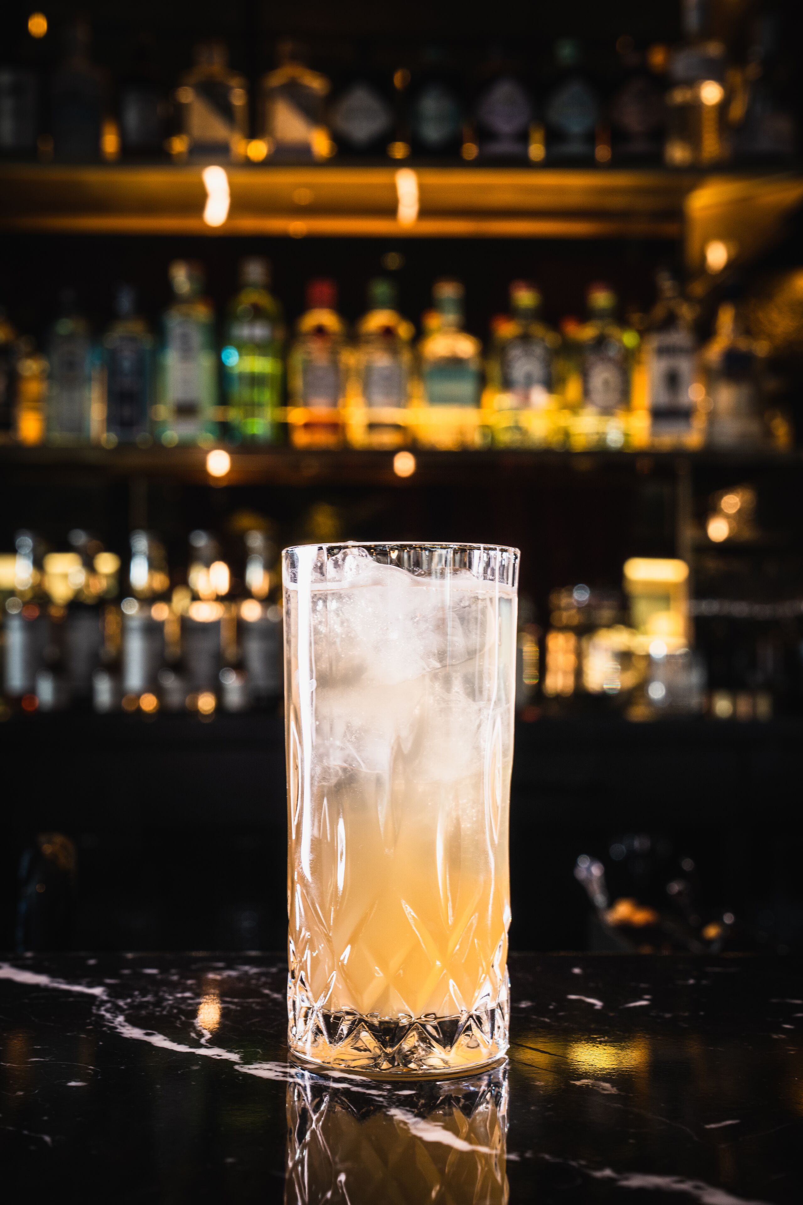 Eine Bar mit einem hohen Glas mit einem klaren, bernsteinfarbenen Getränk, Eiswürfeln und verschwommenen Regalen voller Flaschen im Hintergrund.