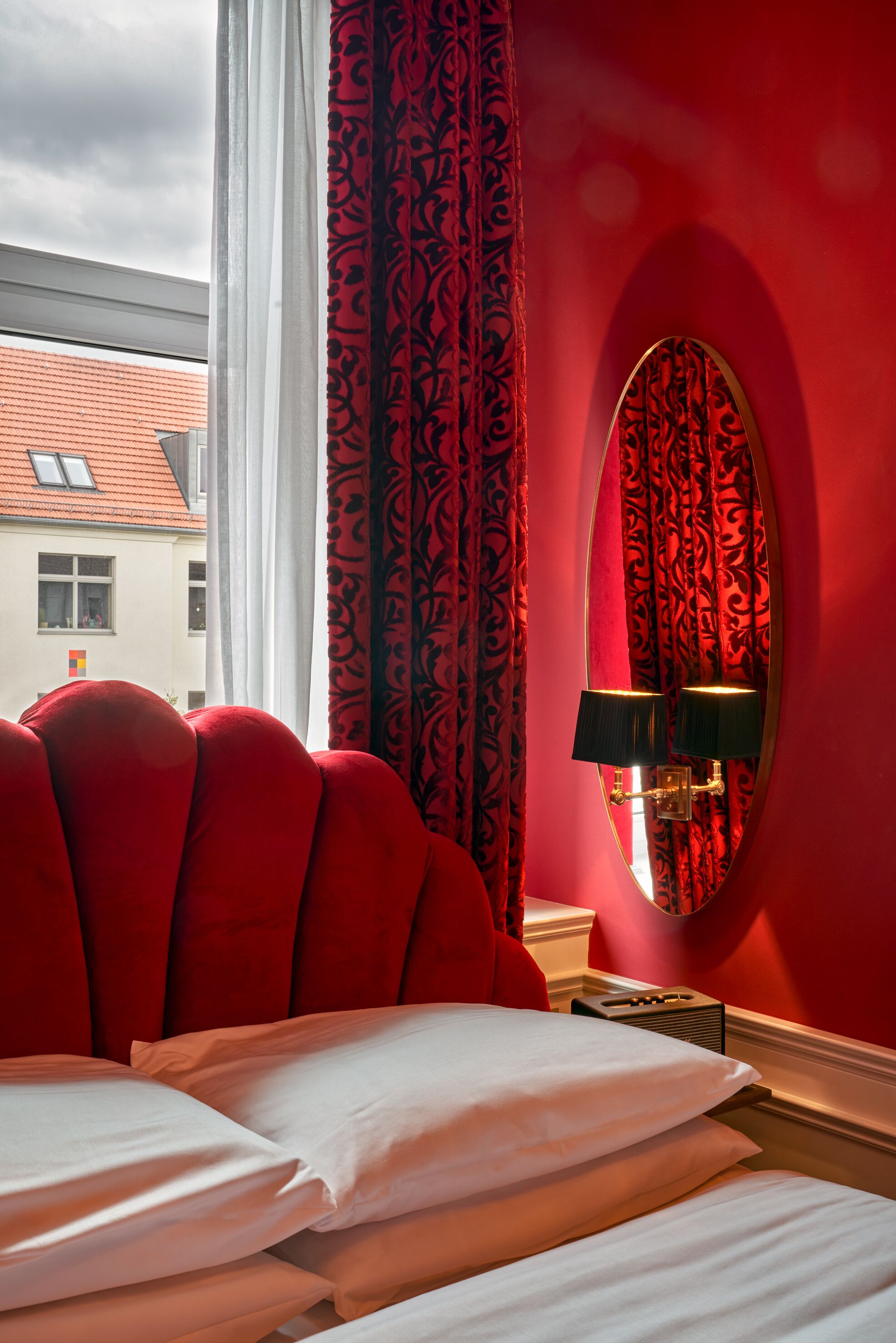 Een hotelkamer met een rode wand, een grote spiegel, een bed met witte lakens en een raam met gordijnen.