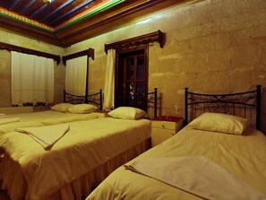 A room with three beds, each with a wooden headboard and white bedding. There is a window with curtains, and the room has a wooden ceiling with a decorative pattern.