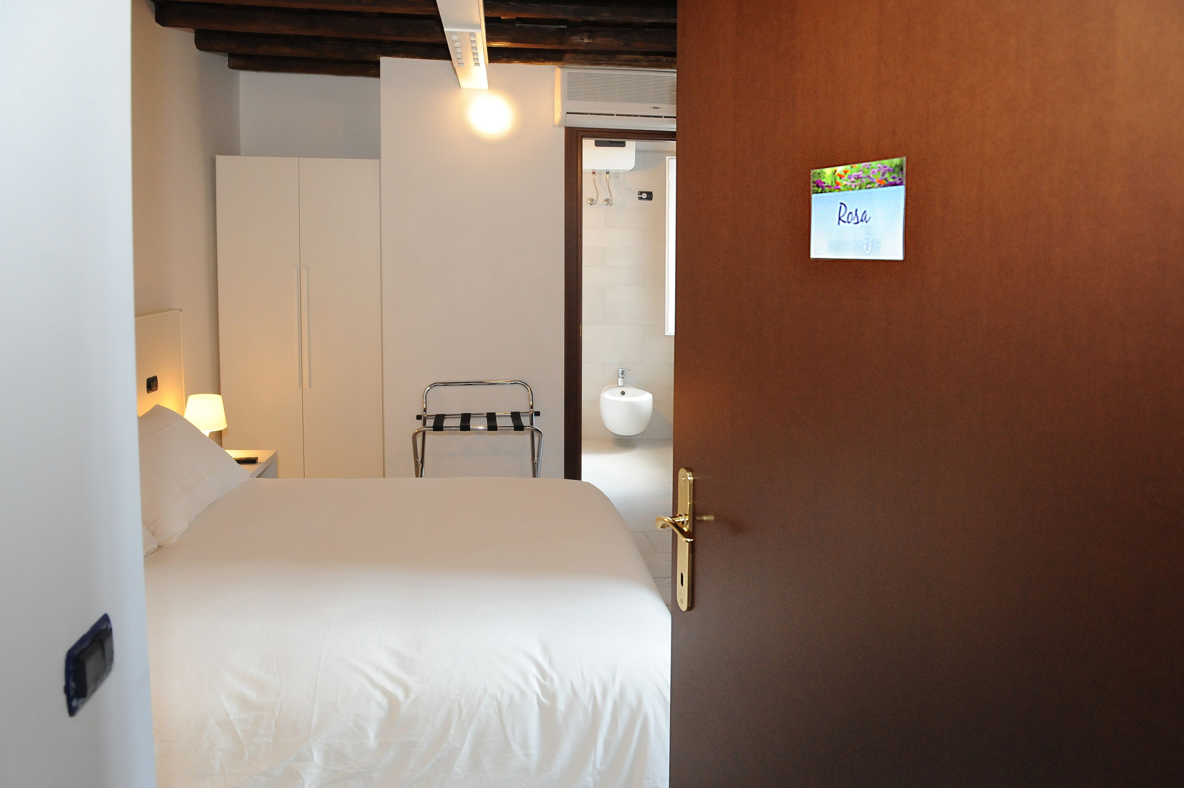 Habitación de hotel con una cama, una mesita de noche con lámpara, un armario, un perchero y un baño visible a través de una puerta abierta.