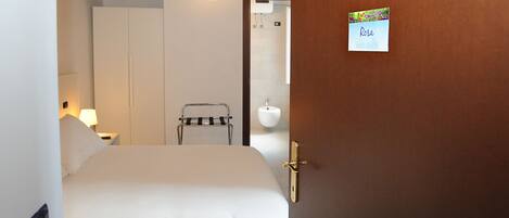 Habitación de hotel con una cama, una mesita de noche con lámpara, un armario, un perchero y un baño visible a través de una puerta abierta.
