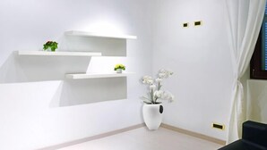Una habitación moderna con paredes blancas, techo de madera y sofá negro. Se observan estanterías blancas con plantas y un jarrón blanco con flores.