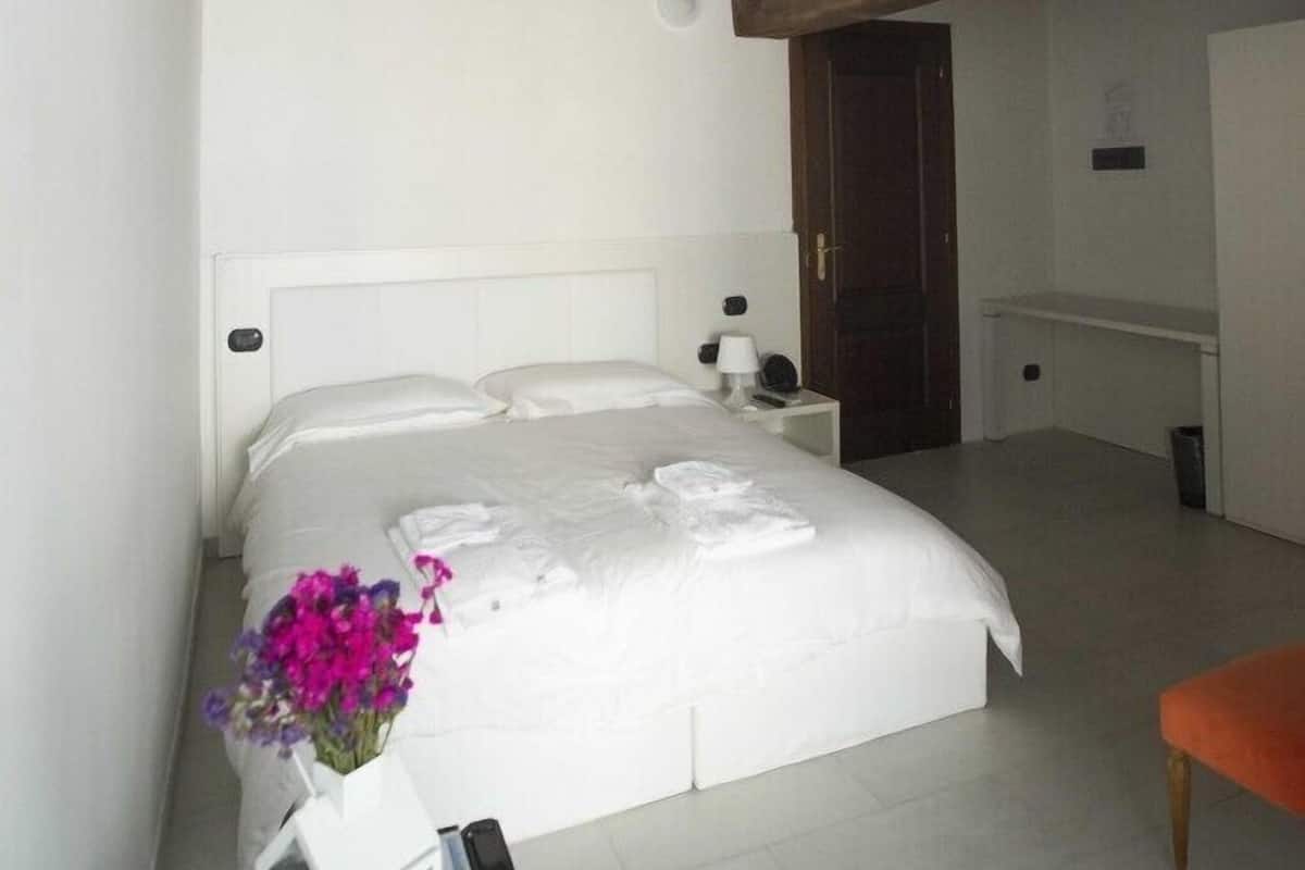 A bedroom with a wooden ceiling, a white bed, a bedside table with a vase of flowers, and a small orange bench.