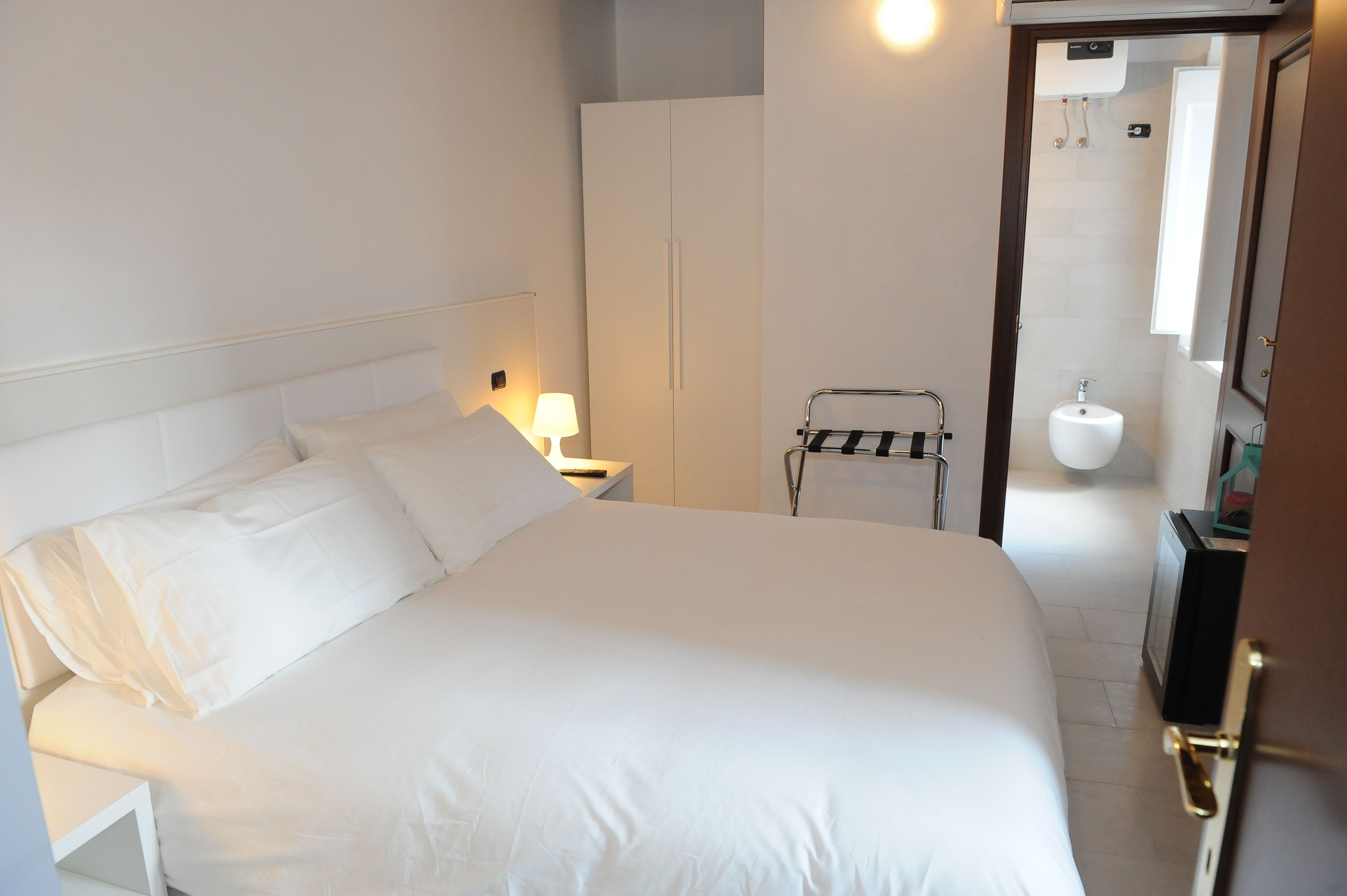 Ein Hotelzimmer mit einem großen Bett, einem Nachttisch mit Lampe, einem Kleiderschrank und einem durchgegangene Tür erkennbaren Badezimmer.