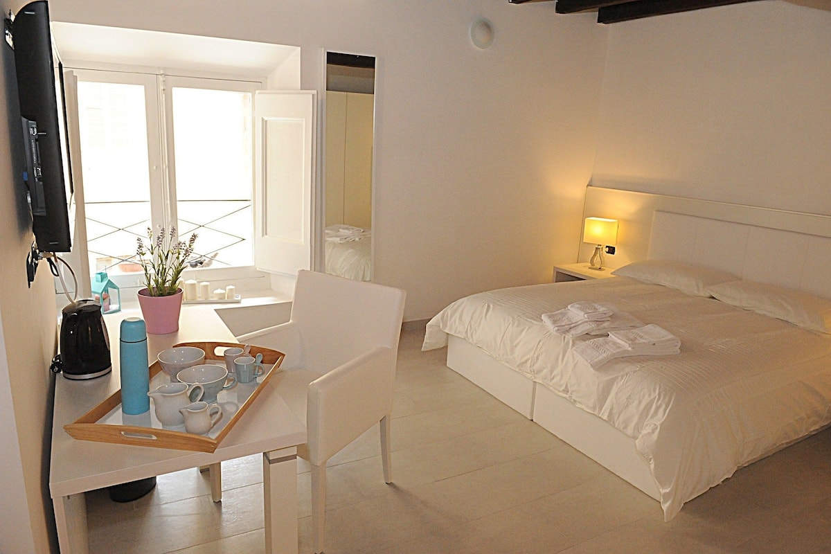 A hotel room with a bed, a desk with a tray of items, a chair, a TV, and a window with a view of a balcony.
