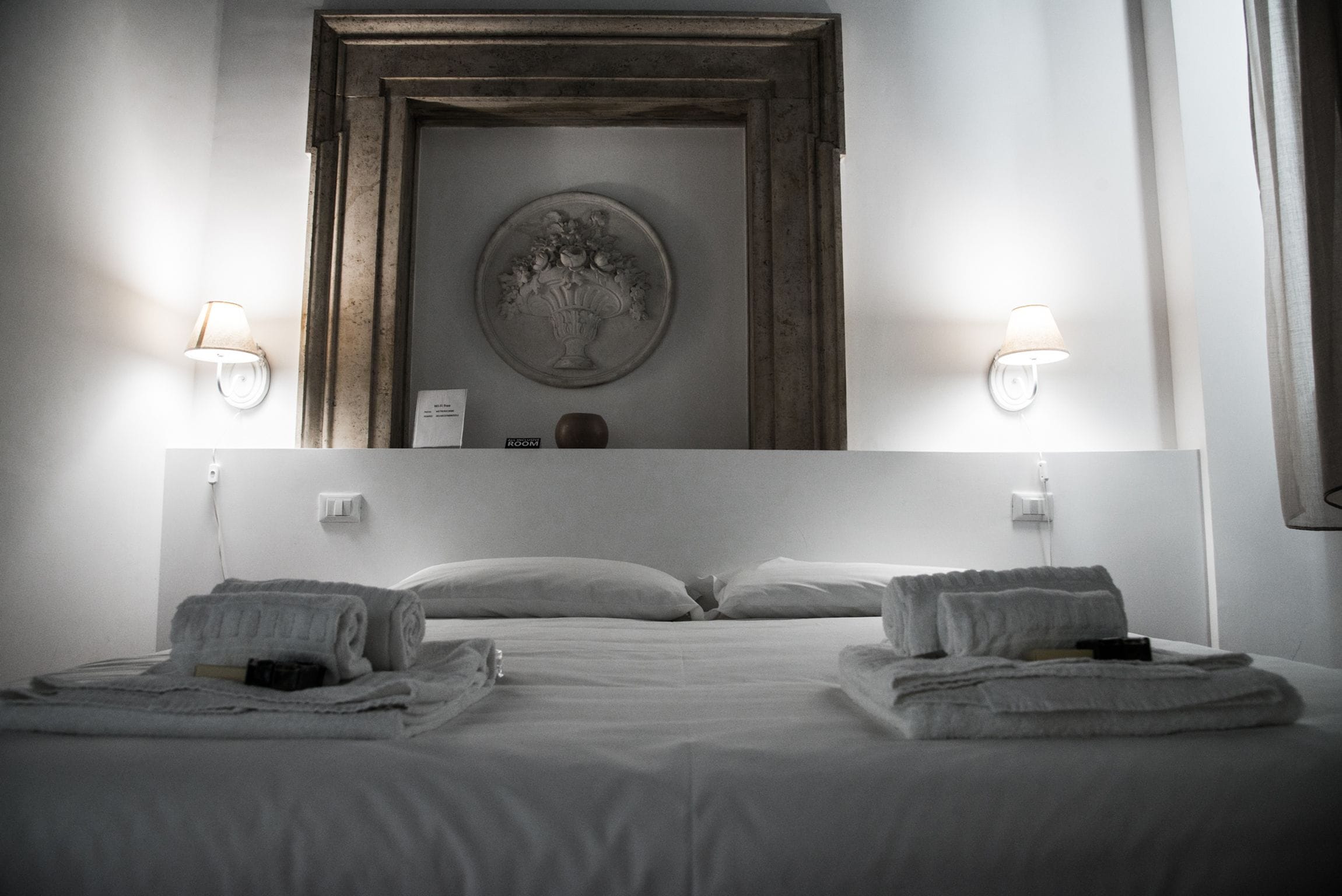 A hotel room with a large bed, two bedside lamps, a mirror above the bed, and a framed artwork on the wall.