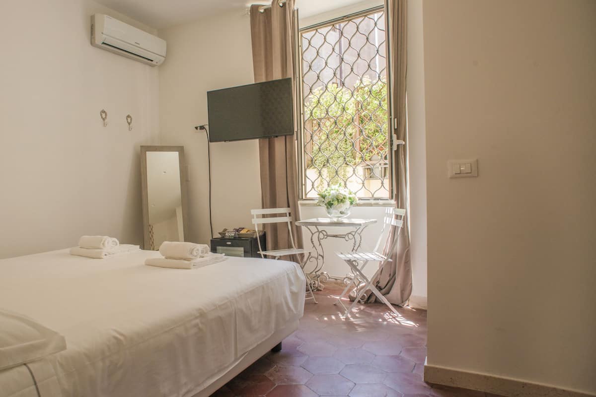 A hotel room with a bed, a desk with a chair, a window with curtains, and an air conditioning unit.