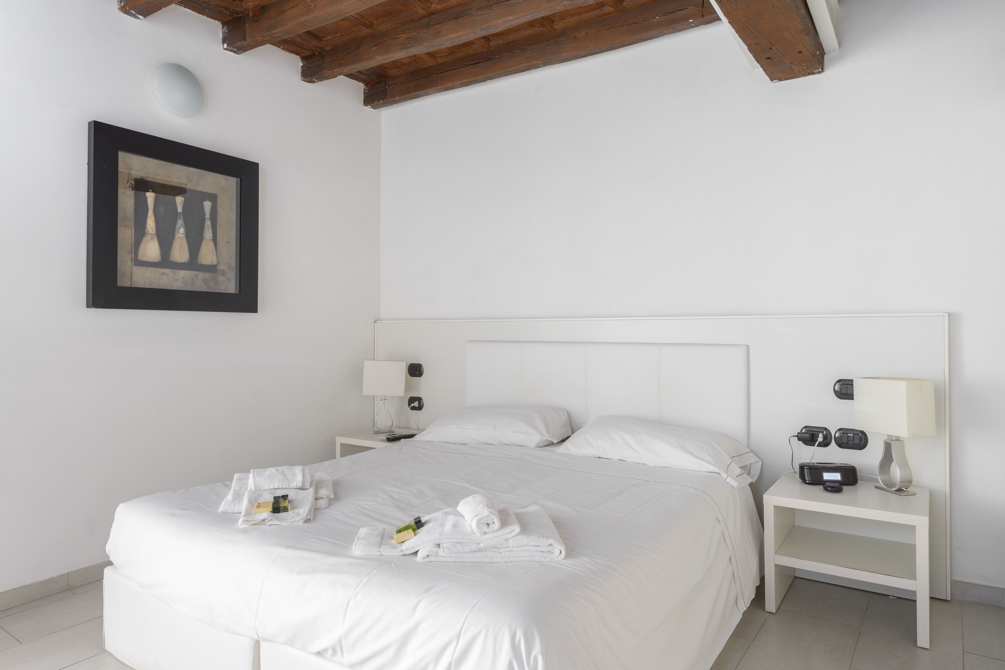 Un lit bien fait, avec du linge de lit et des serviettes blanches, une table de chevet avec une lampe et une radio, et un tableau encadré au mur.