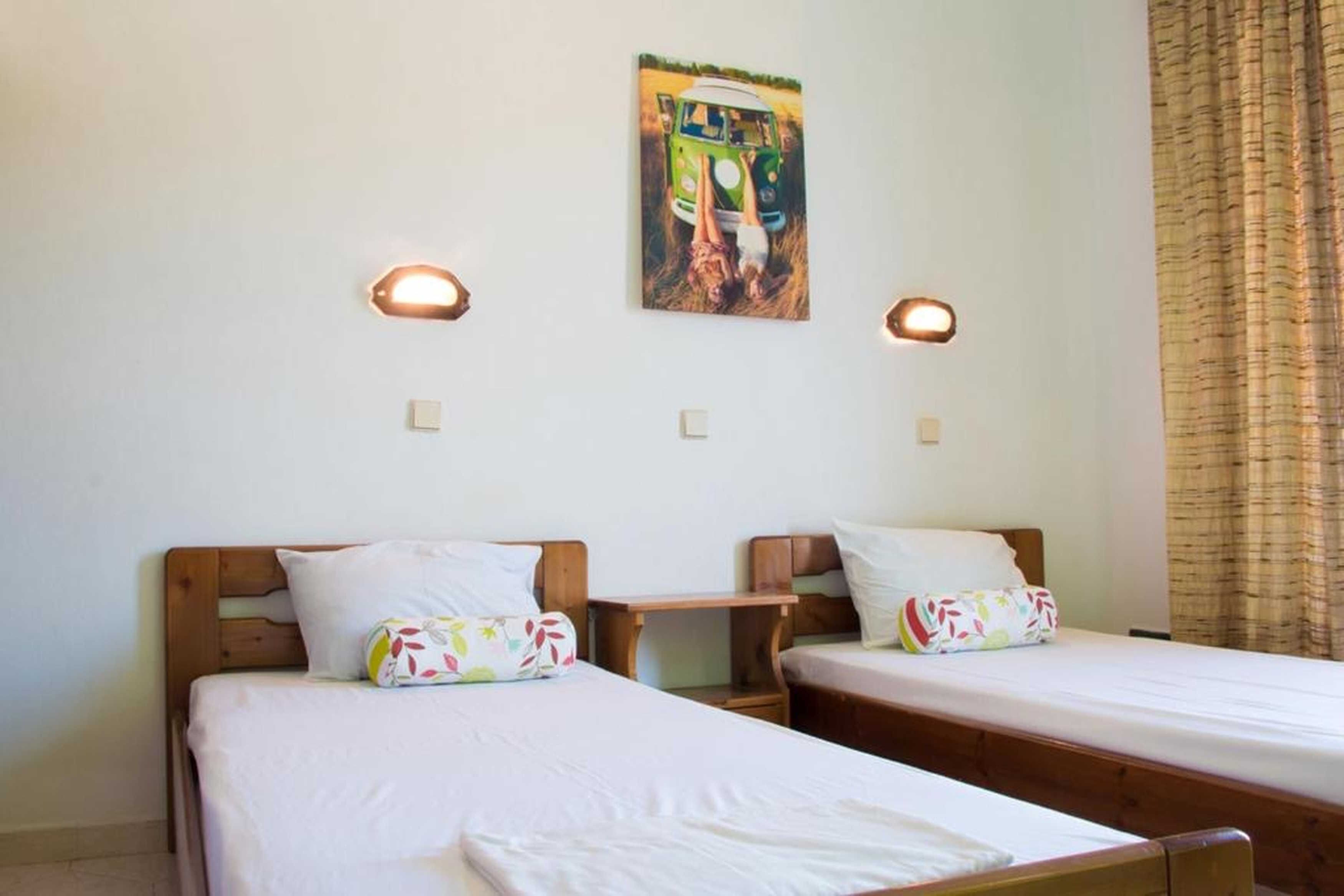 A room with two single beds, each with a wooden headboard, white bedding, and colorful pillows. There are two wall-mounted light fixtures and a painting of a green van on the wall.