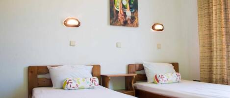 A room with two single beds, each with a wooden headboard, white bedding, and colorful pillows. There are two wall-mounted light fixtures and a painting of a green van on the wall.