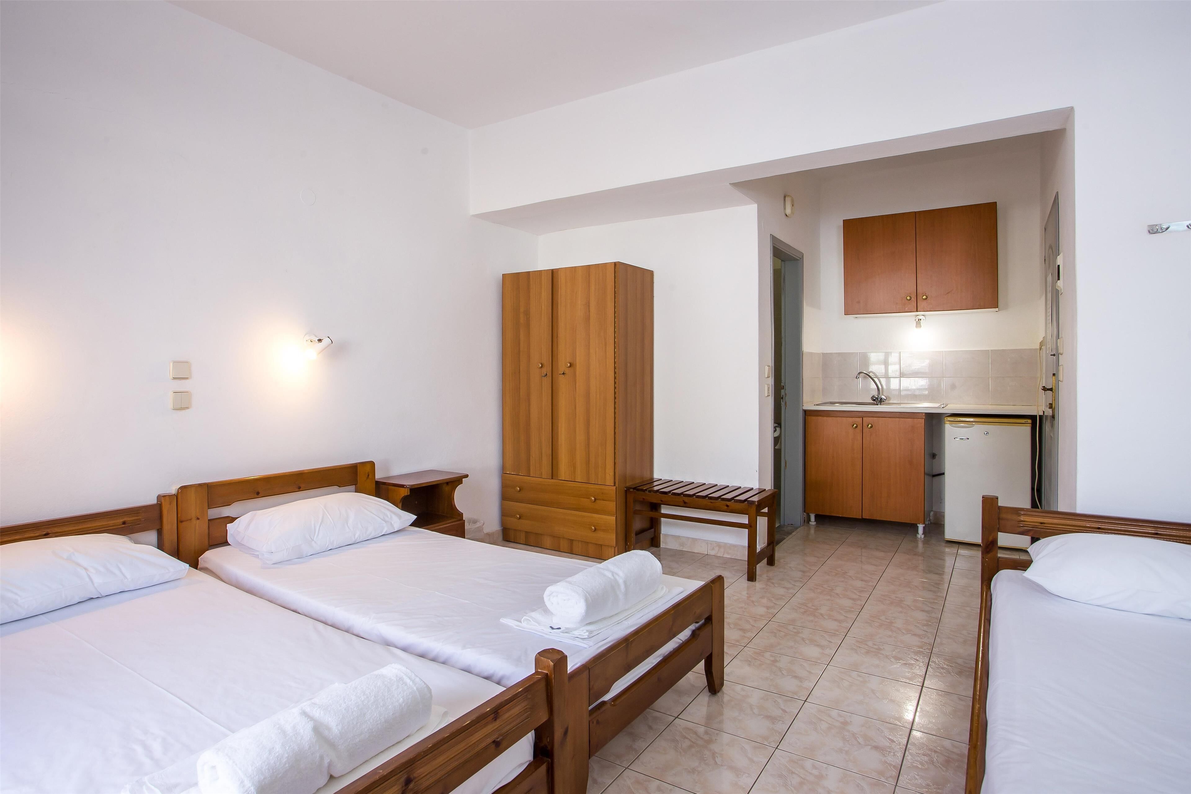 A bedroom with two beds, a wooden wardrobe, a small table, and a kitchenette in the background.
