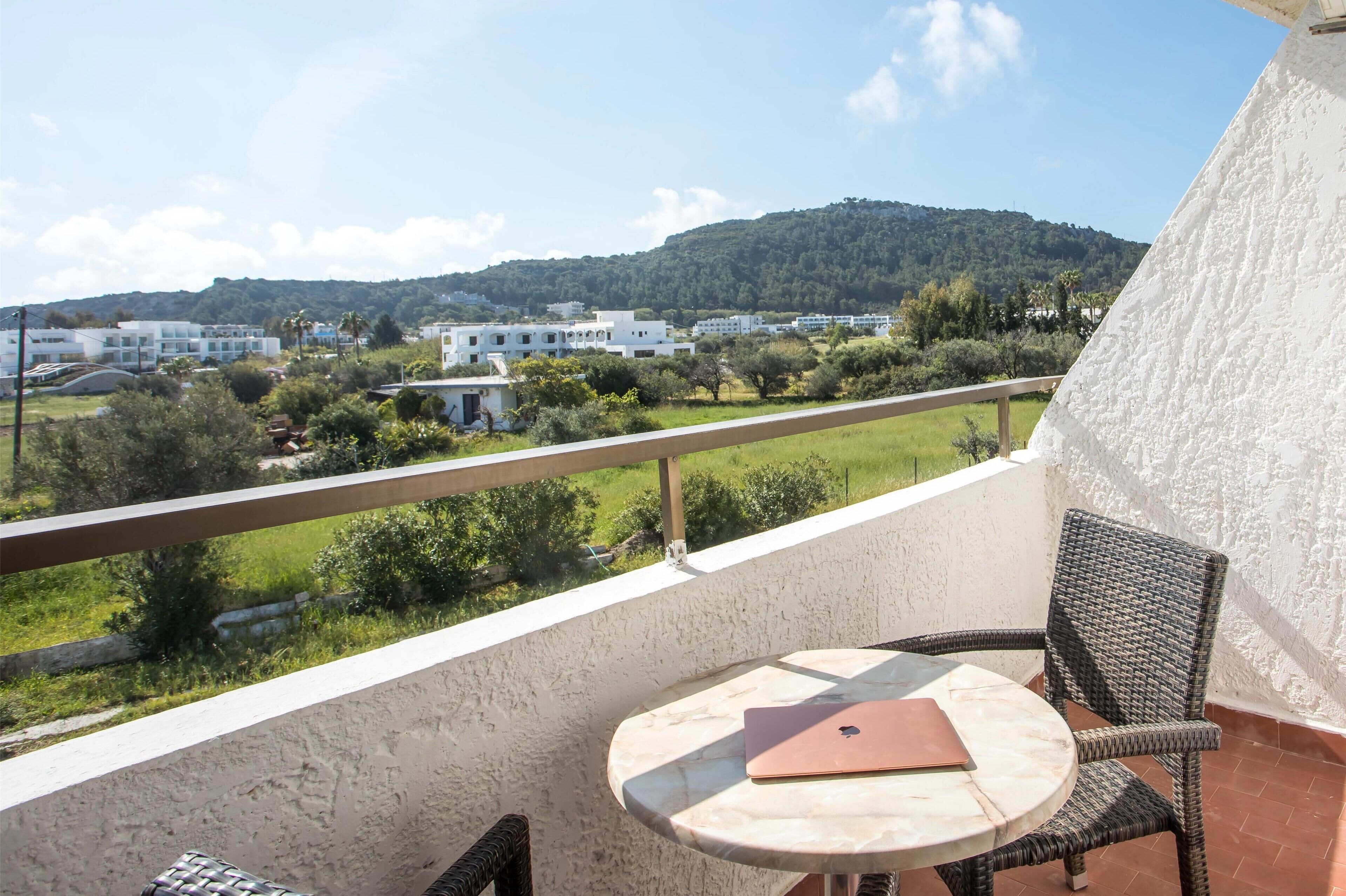 A balcony with a table and chair, overlooking a green landscape and buildings in the distance.