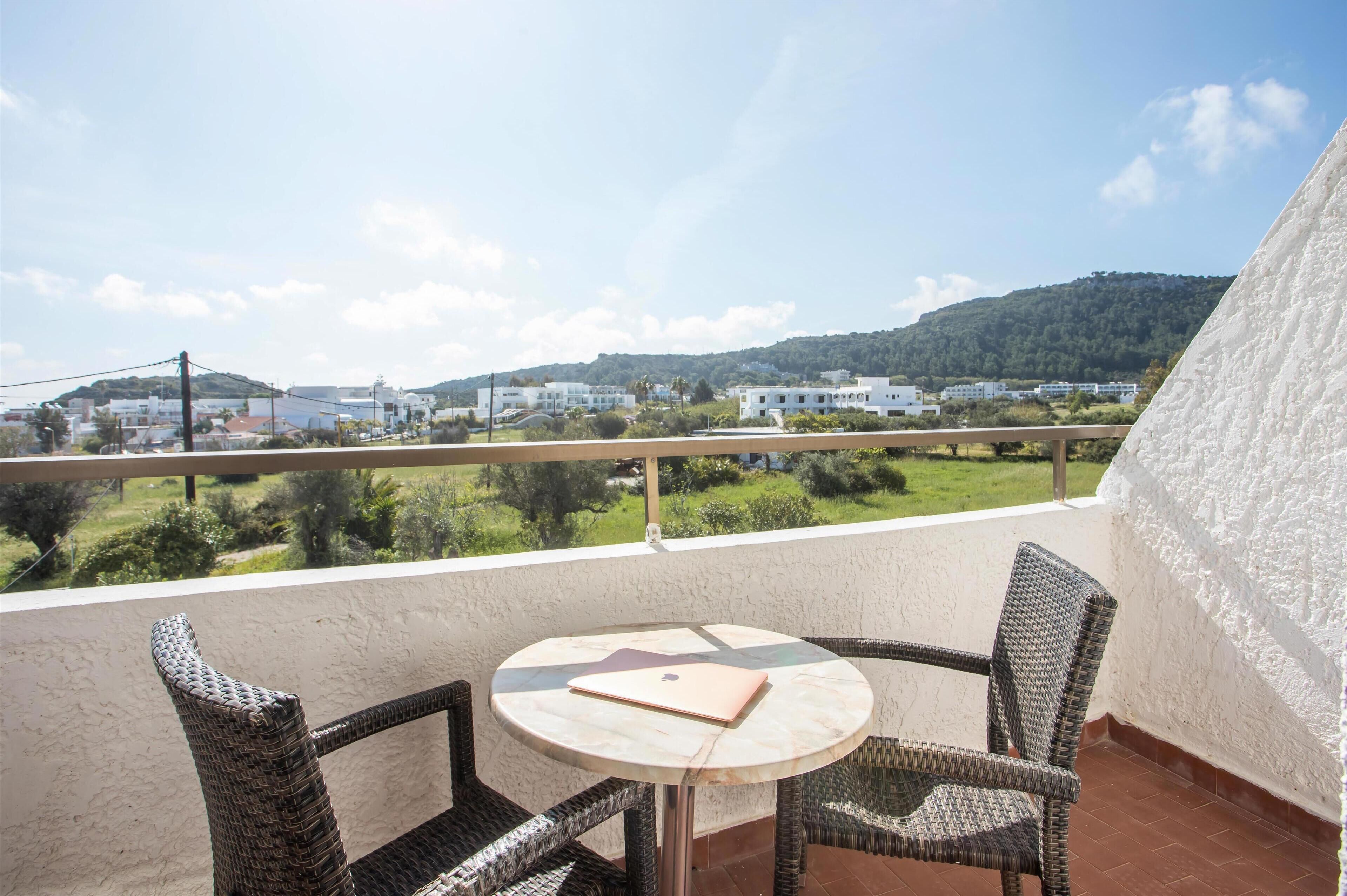 A balcony with wicker chairs and a table, overlooking a green landscape and buildings.