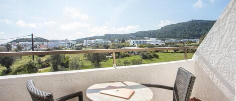 A balcony with wicker chairs and a table, overlooking a green landscape and buildings.