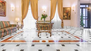 A lobby with a patterned floor, a sofa, a table with a vase, and curtains. - Raeli Hotel Noto (Rome)