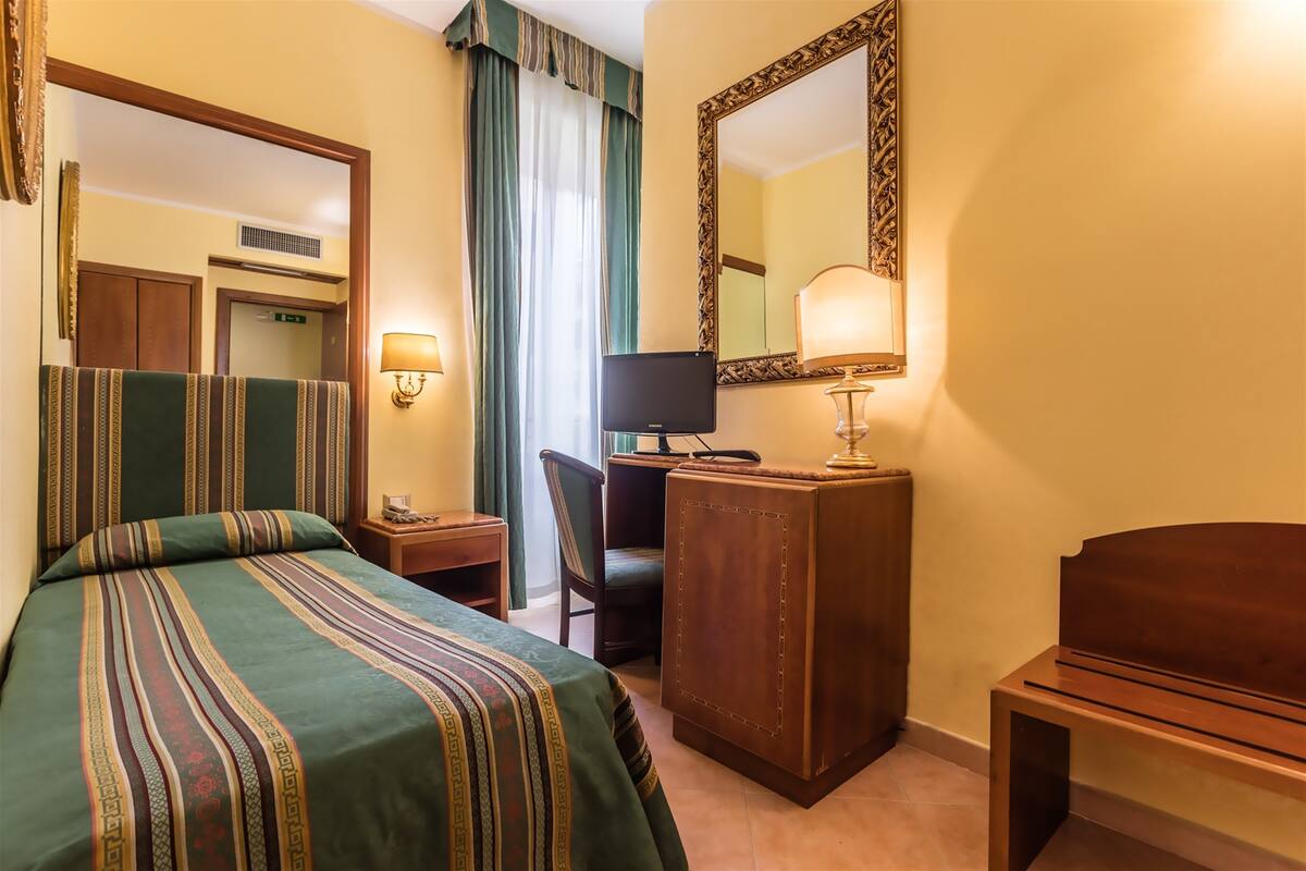 Camera d'albergo con un letto, una scrivania, una sedia, una televisione, uno specchio e una lampada.