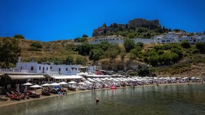 Eine Küstenstadt mit einem Schloss auf einem Hügel, einem Strand mit Sonnenschirmen und einem Gasthof namens "Taverna Skala".