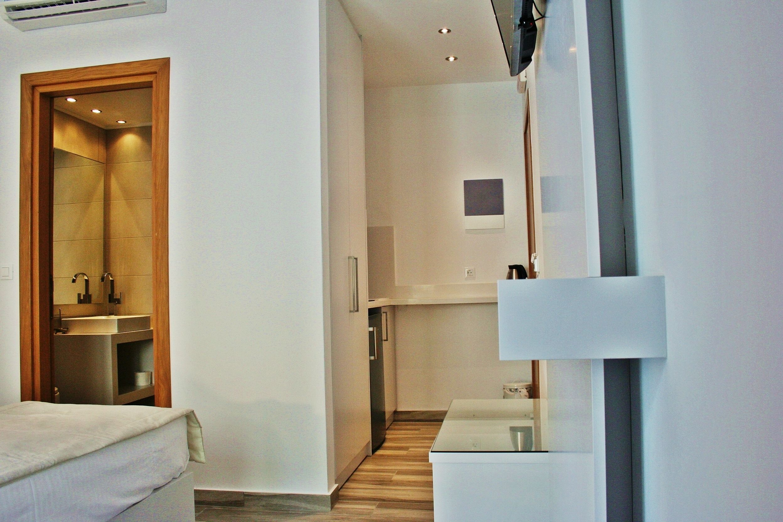 A modern hotel room with a wooden door leading to a bathroom, a small kitchen area, and a bed with a white headboard.
