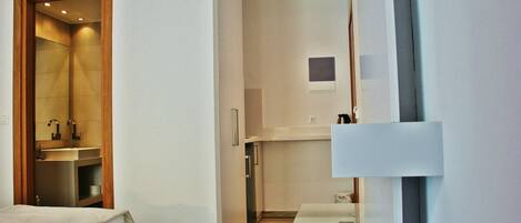 A modern hotel room with a wooden door leading to a bathroom, a small kitchen area, and a bed with a white headboard.