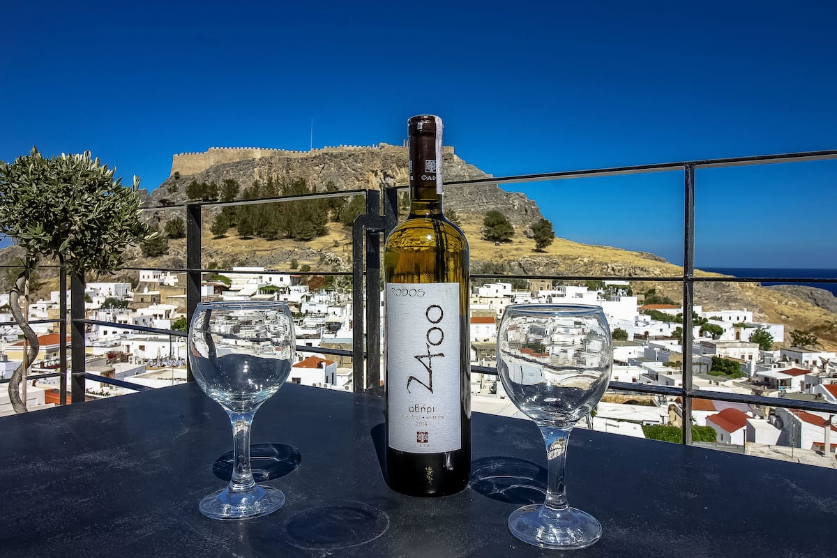 A bottle of wine with a label, two empty wine glasses, and a scenic view of a coastal town with a castle in the background.