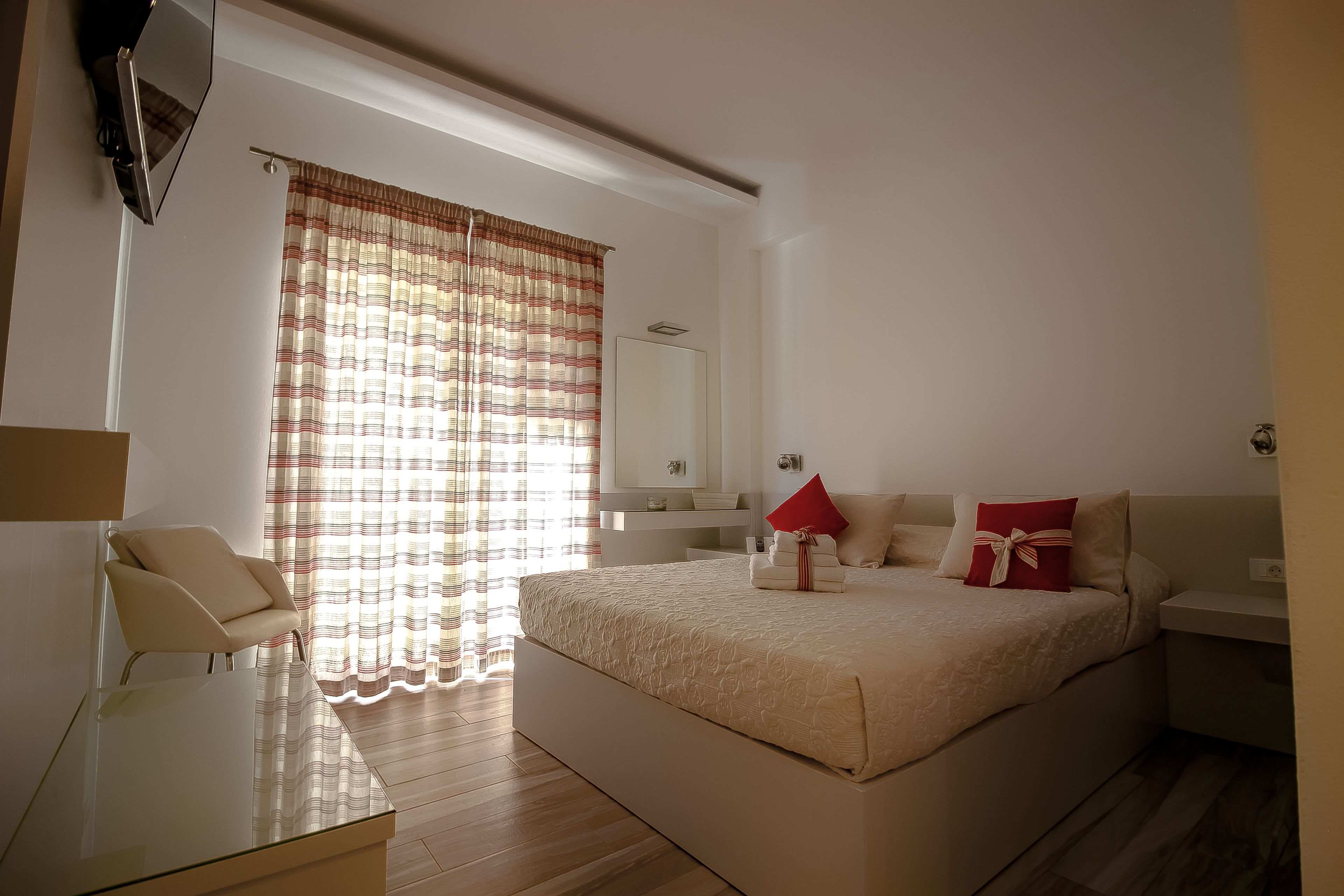 Ein Schlafzimmer mit einem großen Bett, einem Nachttisch, einem Sessel, einem Spiegel und einem Fenster mit Vorhängen.