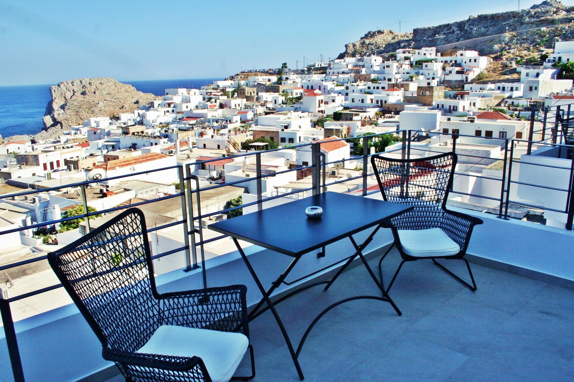 A balcony with two chairs and a table, overlooking a coastal town with white buildings and a rocky hill.