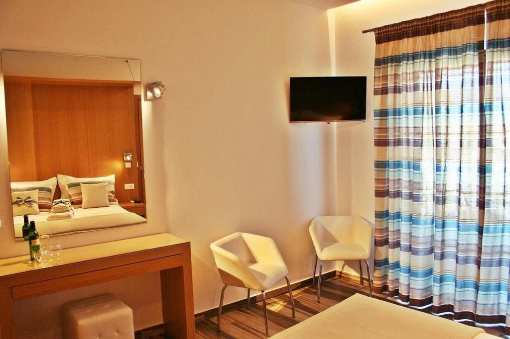 A hotel room with a bed, a desk with a mirror, two white chairs, a flat-screen TV mounted on the wall, and striped curtains.