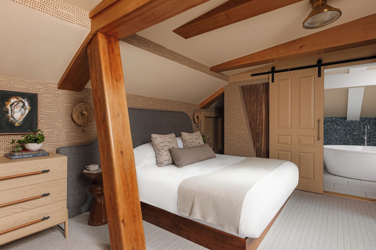 Une chambre avec un plafond en bois, un grand lit, une commode, une petite table avec une plante, et une salle de bain avec une baignoire.
