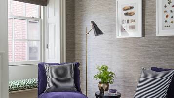 A room with two purple velvet armchairs, a grey ottoman, a floor lamp, and a window with a view of a brick building.